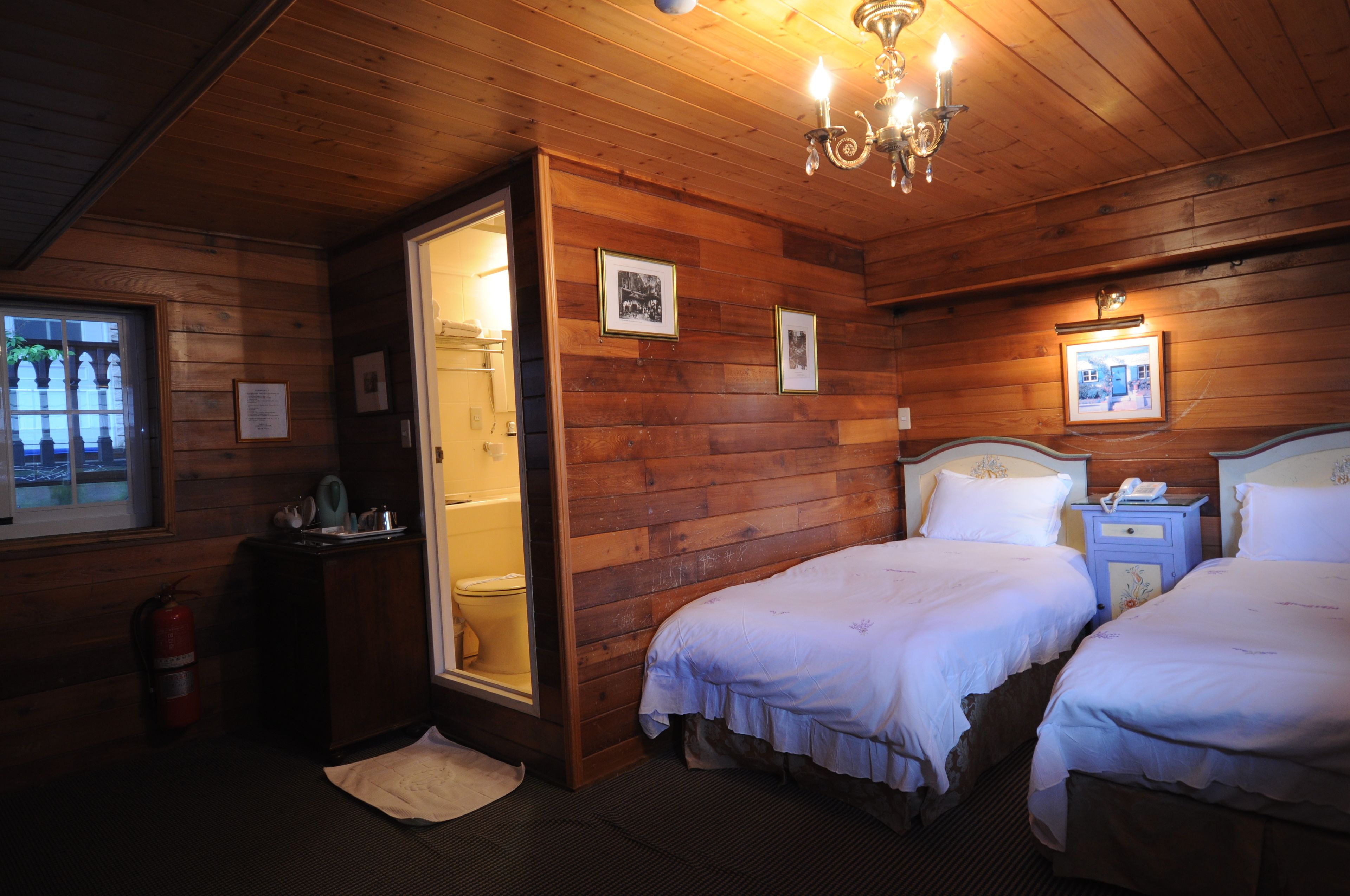 A wooden-paneled room with two beds, a bedside table with a phone, and a visible toilet.