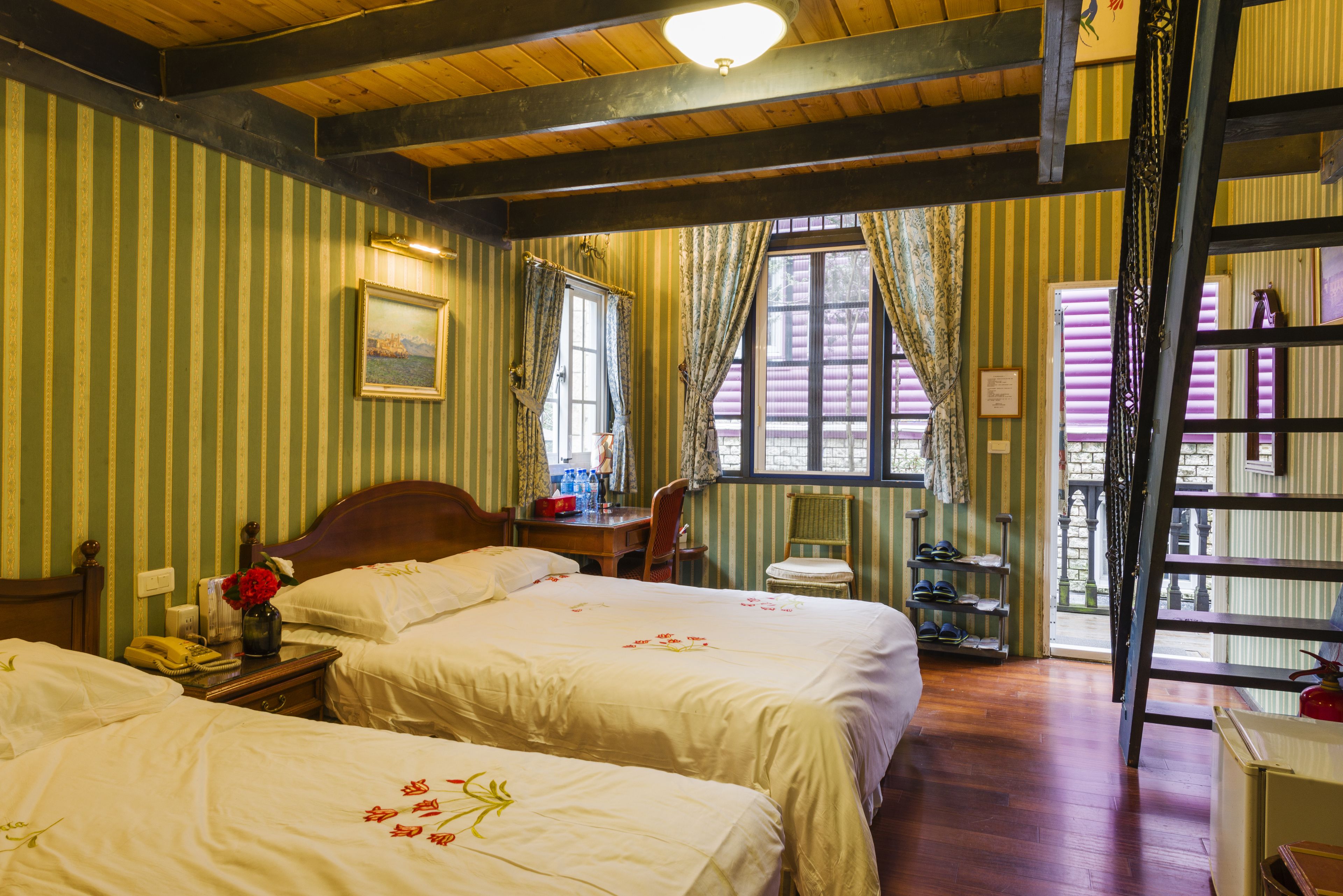 Una habitación con dos camas, un escritorio de madera, una silla, un ventanal con cortinas y una escalera que lleva a un balcón.