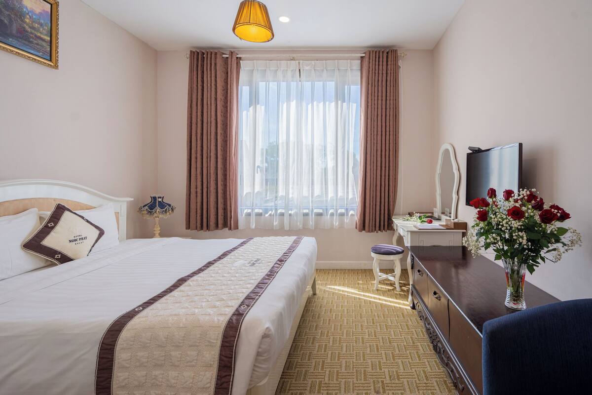 Une chambre d’hôtel avec un lit, un bureau, une chaise, une télévision et un vase avec des fleurs.