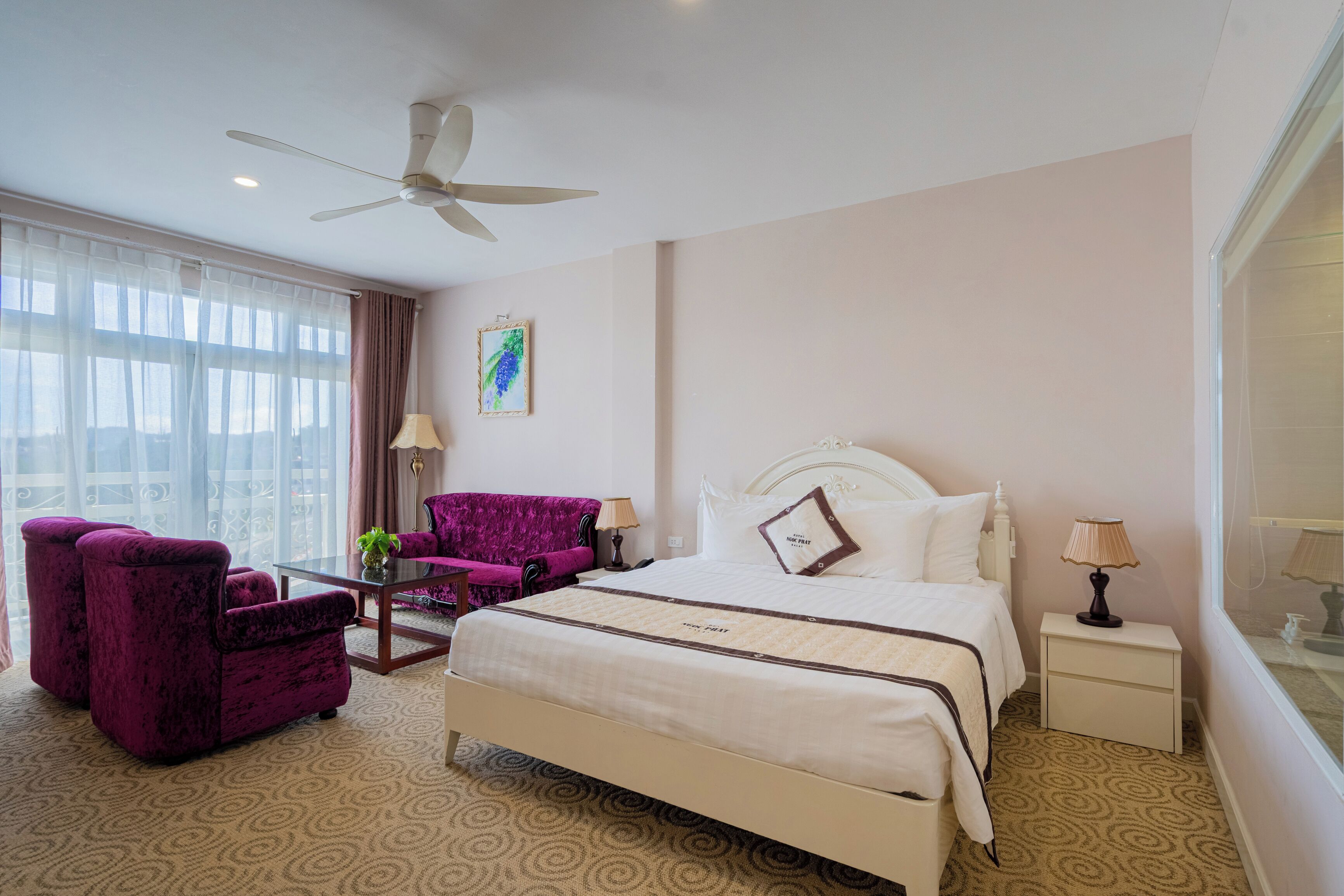 A hotel room with a bed, a purple armchair, a small table, a ceiling fan, and a window with curtains.