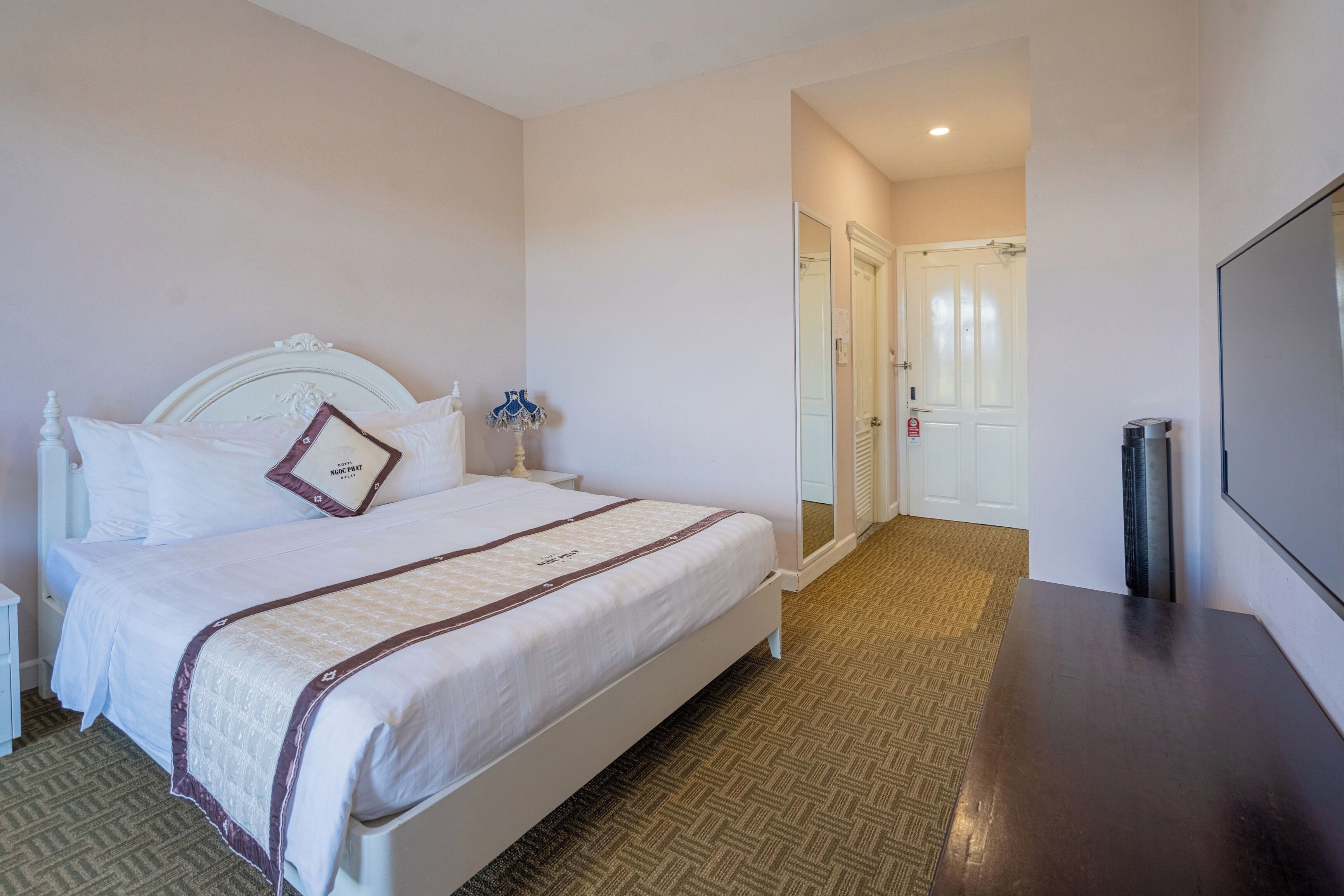 Habitación de hotel con una cama bien hecha, una mesita de noche con lámpara, un espejo, una puerta que da a otra habitación y un ventanal grande.