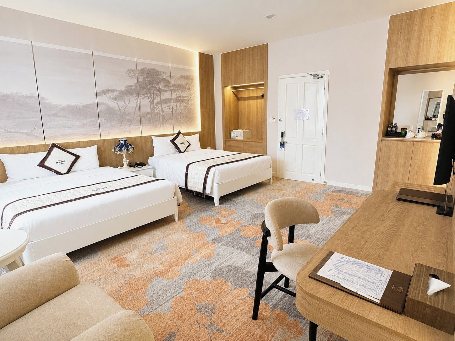 A hotel room with two beds, a desk, a chair, a TV, and a door.
