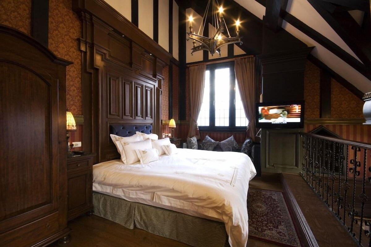 A traditional room with a large bed, wooden paneling, a chandelier, and a view of the outside through a window.