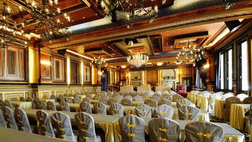 A banquet hall with rows of tables and chairs, ornate chandeliers, and a fireplace.