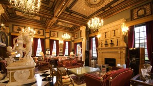 Un lobby de hotel lujoso con candelabros ornamentados, una chimenea imponente, asientos cómodos y un suelo a cuadros.