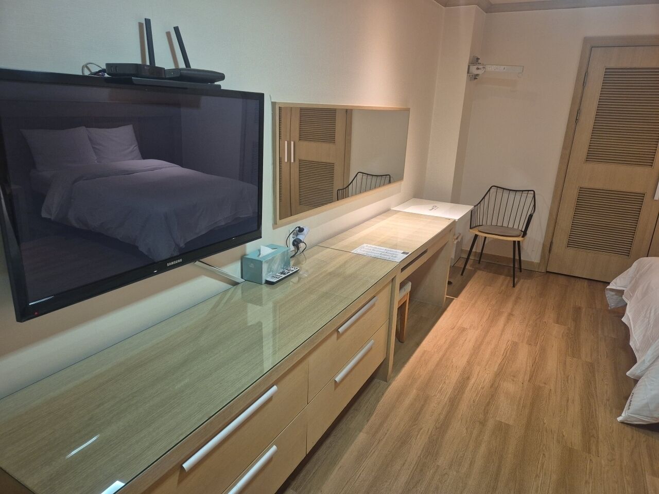A modern hotel room with a large flat-screen TV, a wooden desk, a chair, and a bed.