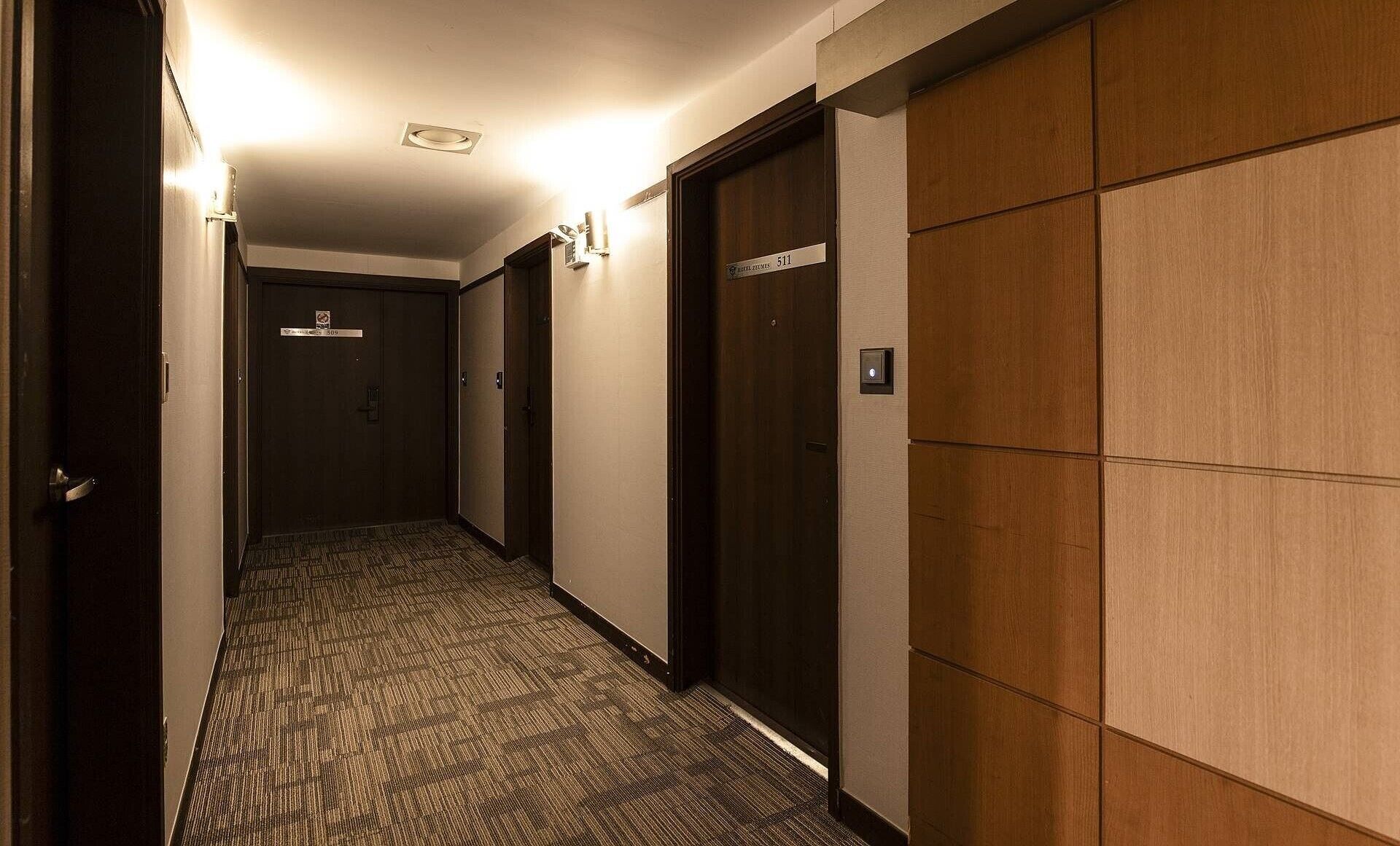 Et hotel med et tæppe belagt gulv, trædøre og et elevatorstyrepaneel på væggen.