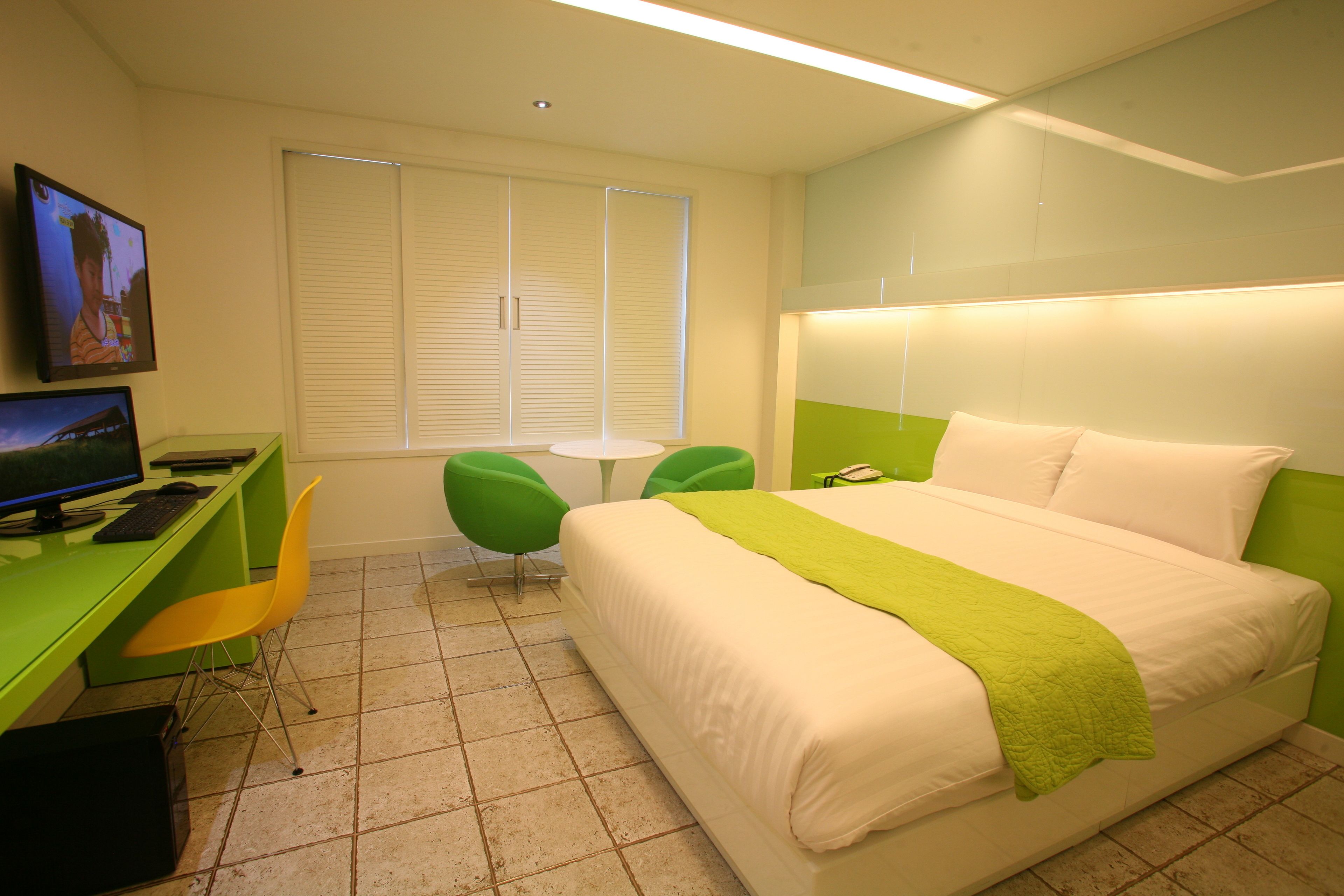Una habitación de hotel con cama, escritorio con computadora, dos sillas, una mesita y televisor en la pared.