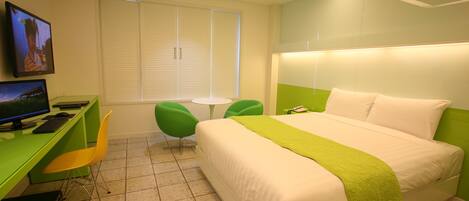 Una habitación de hotel con cama, escritorio con computadora, dos sillas, una mesita y televisor en la pared.