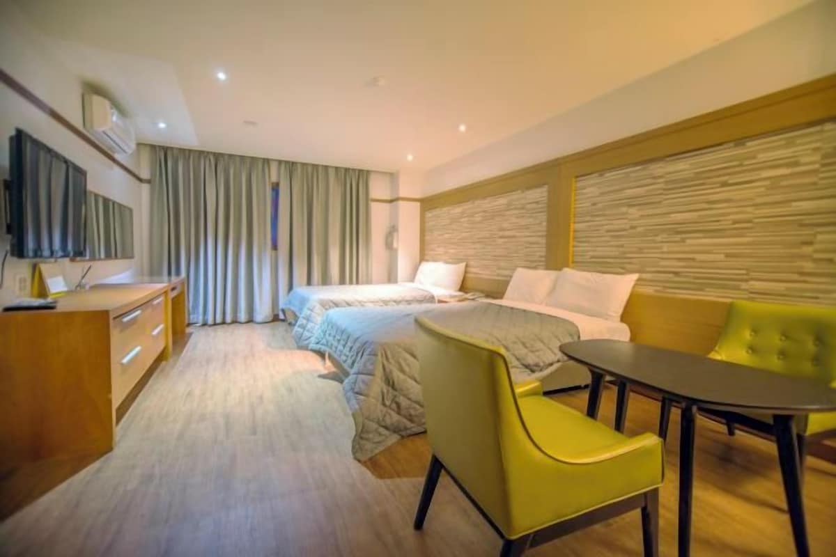 A modern hotel room with a wooden desk, a bed with two pillows, a sofa, and a TV mounted on the wall.