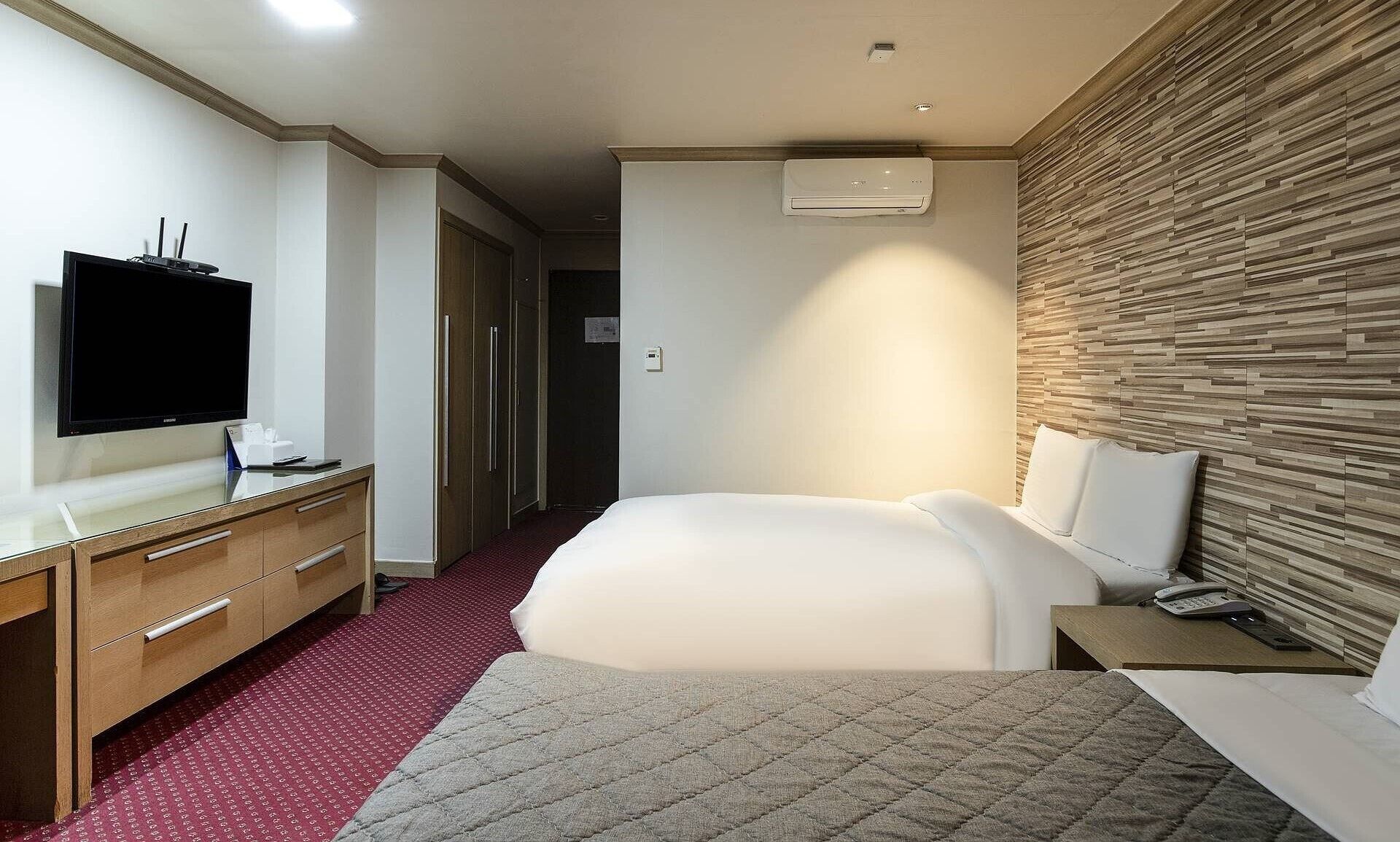 A hotel room with a bed, a desk with a flat-screen TV, a wooden headboard, and a red carpeted floor.