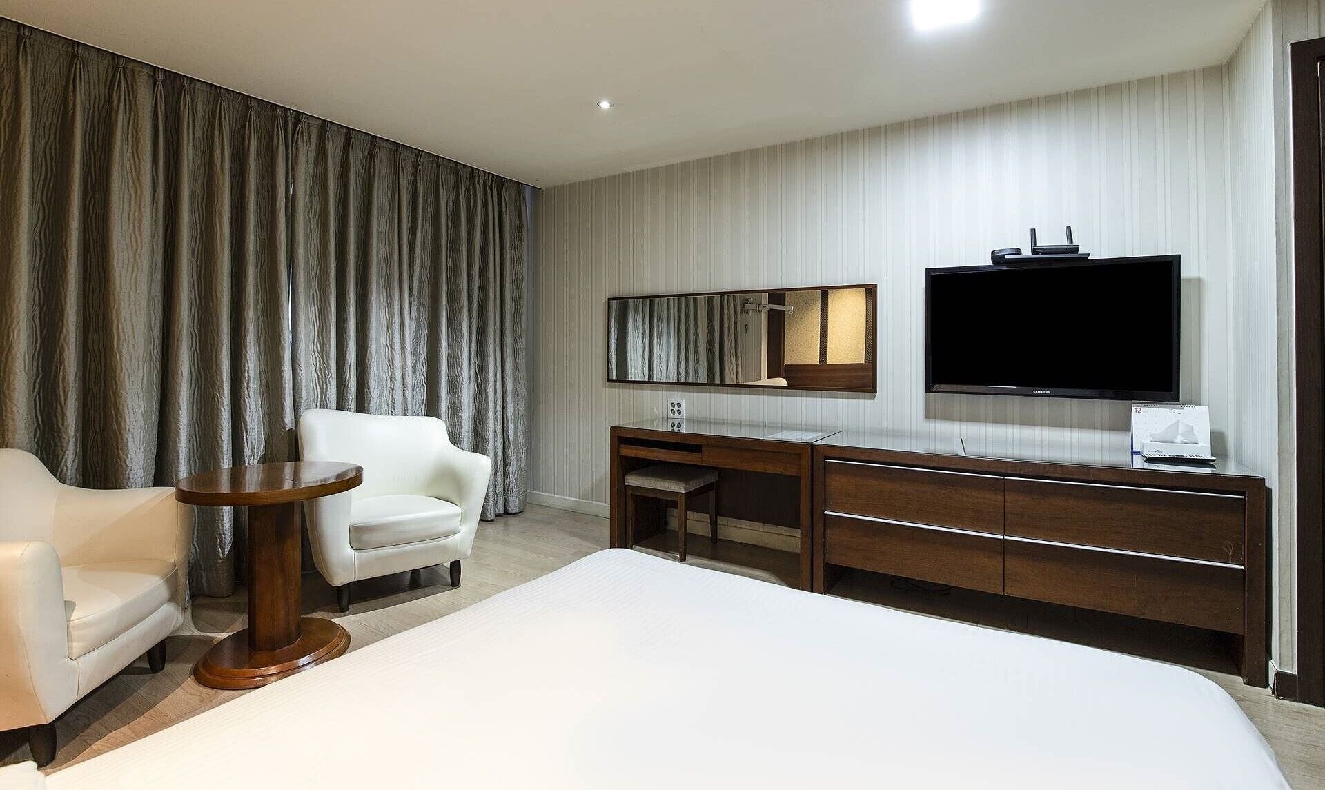 Ein Hotelzimmer mit einem Bett, einem Schreibtisch, einem Sessel, einem Fernseher und einem großen Fenster mit Vorhängen.