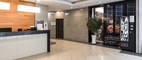 A hotel reception area with a wooden reception desk, a potted plant, a glass door leading to the restaurant, and a vending machine.