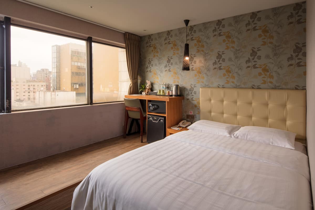 A hotel room with a large bed, a desk with a chair, a small refrigerator, a telephone, and a window with a view of buildings.