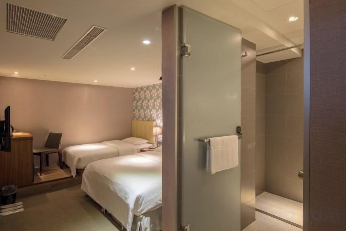 A hotel room with two beds, a desk, a chair, a shower, and a towel rack.