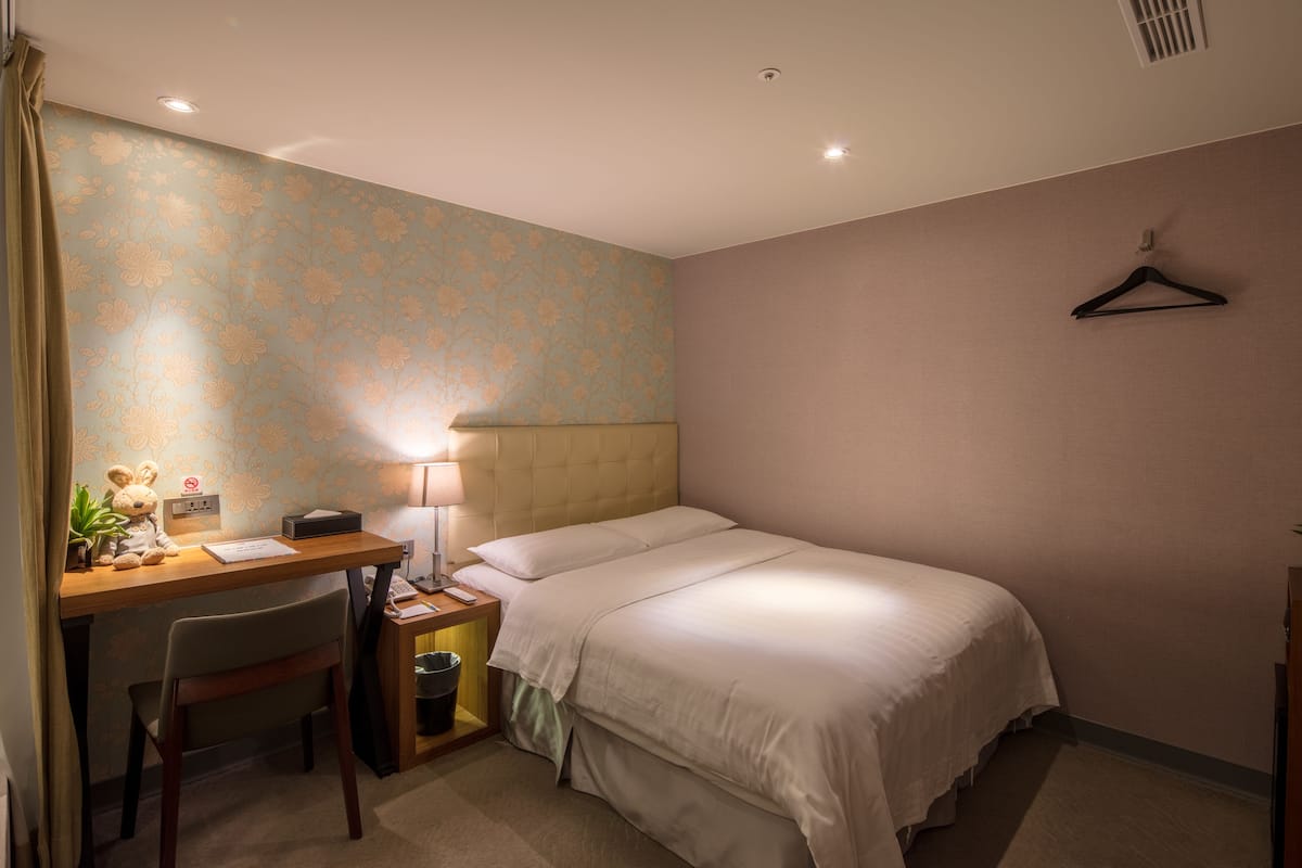 A hotel room with a bed, a desk with a chair, a lamp, and a hanging coat rack.