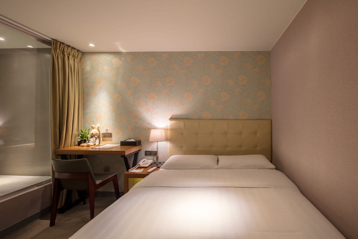 A hotel room with a bed, desk, chair, lamp, and a small table with a plant.