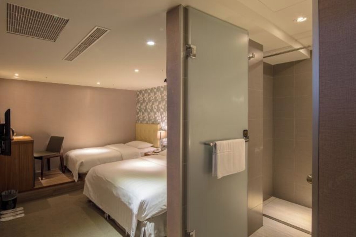 A hotel room with two beds, a desk, a chair, a shower area, and a wall-mounted towel rack.