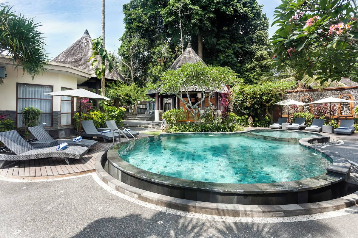 A circular hot tub surrounded by lounge chairs and umbrellas, with a tropical garden and a gazebo in the background.