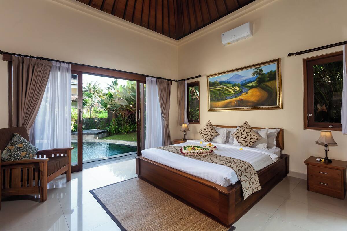 A spacious bedroom with a large bed, a wooden nightstand, a painting, and a view of a pool and garden.