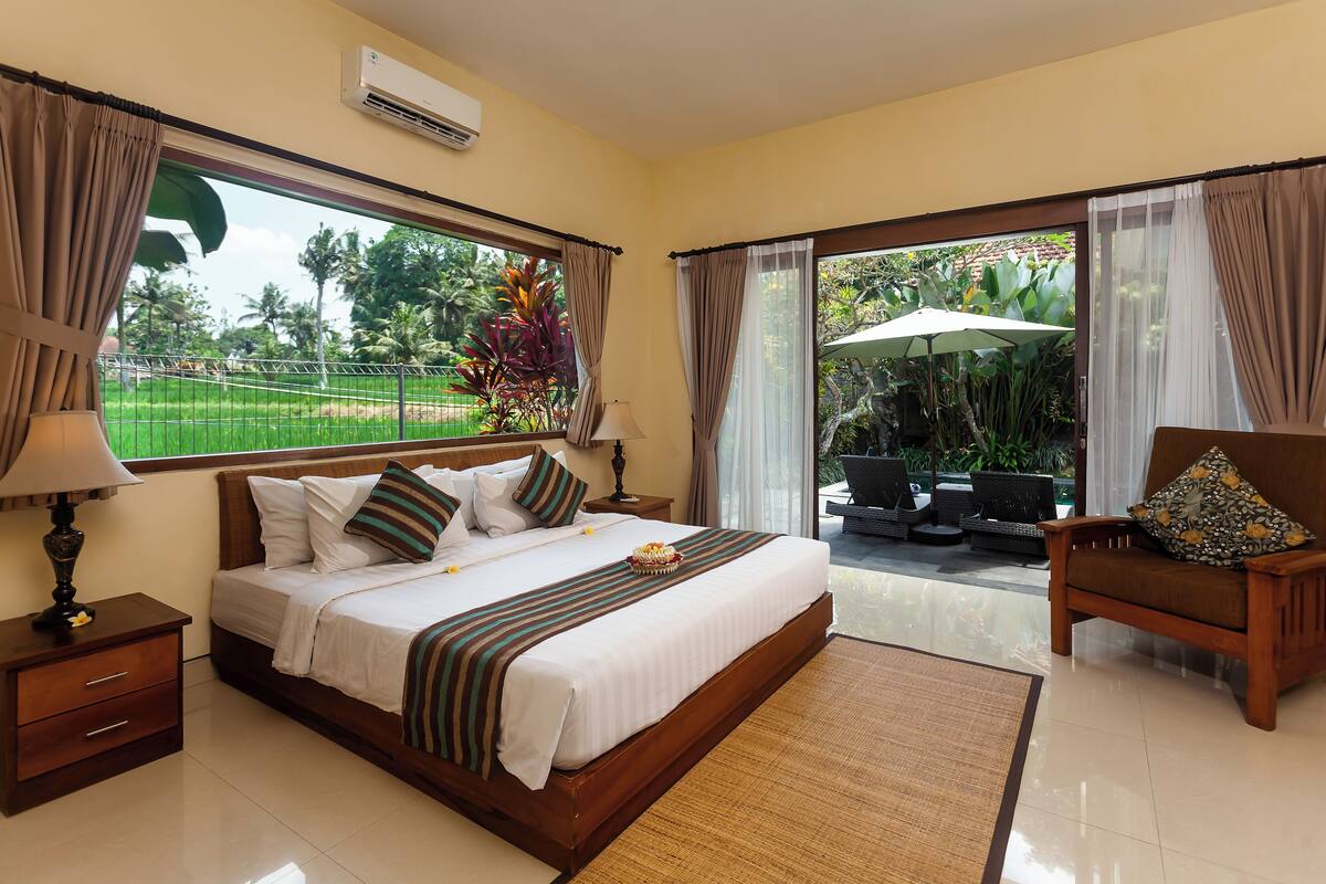 A hotel room with a large bed, a nightstand, a lamp, a view of a garden through a sliding door, and a chair with a floral pillow.