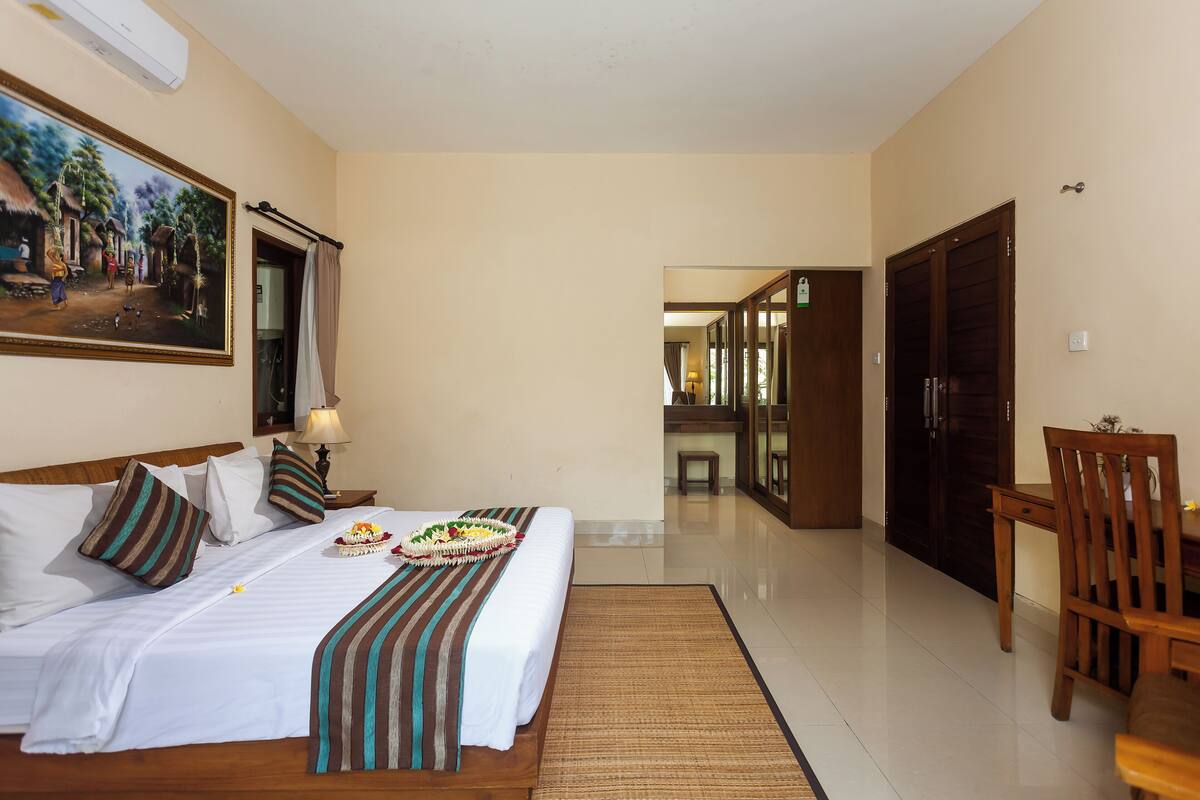 A hotel room with a large bed, a dining table, a chair, a painting on the wall, and a view into the corridor.