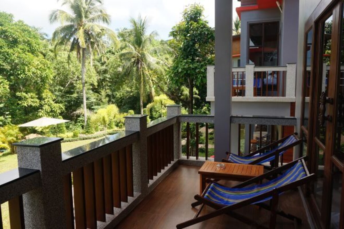 A balcony with a wooden table and striped chairs, overlooking a lush garden with palm trees.