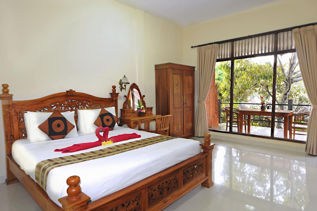 A neatly made bed with a wooden headboard, a wooden nightstand with a lamp, and a view of a balcony with a table and chairs.
