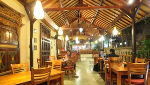 An indoor dining area with wooden tables and chairs, a tiled floor, and a high wooden ceiling with hanging lights. - Rahayu 2 Bungalows (Ubud)