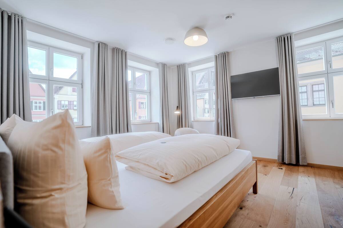 A modern bedroom with a large bed, two windows, a flat-screen TV, and a wooden floor.