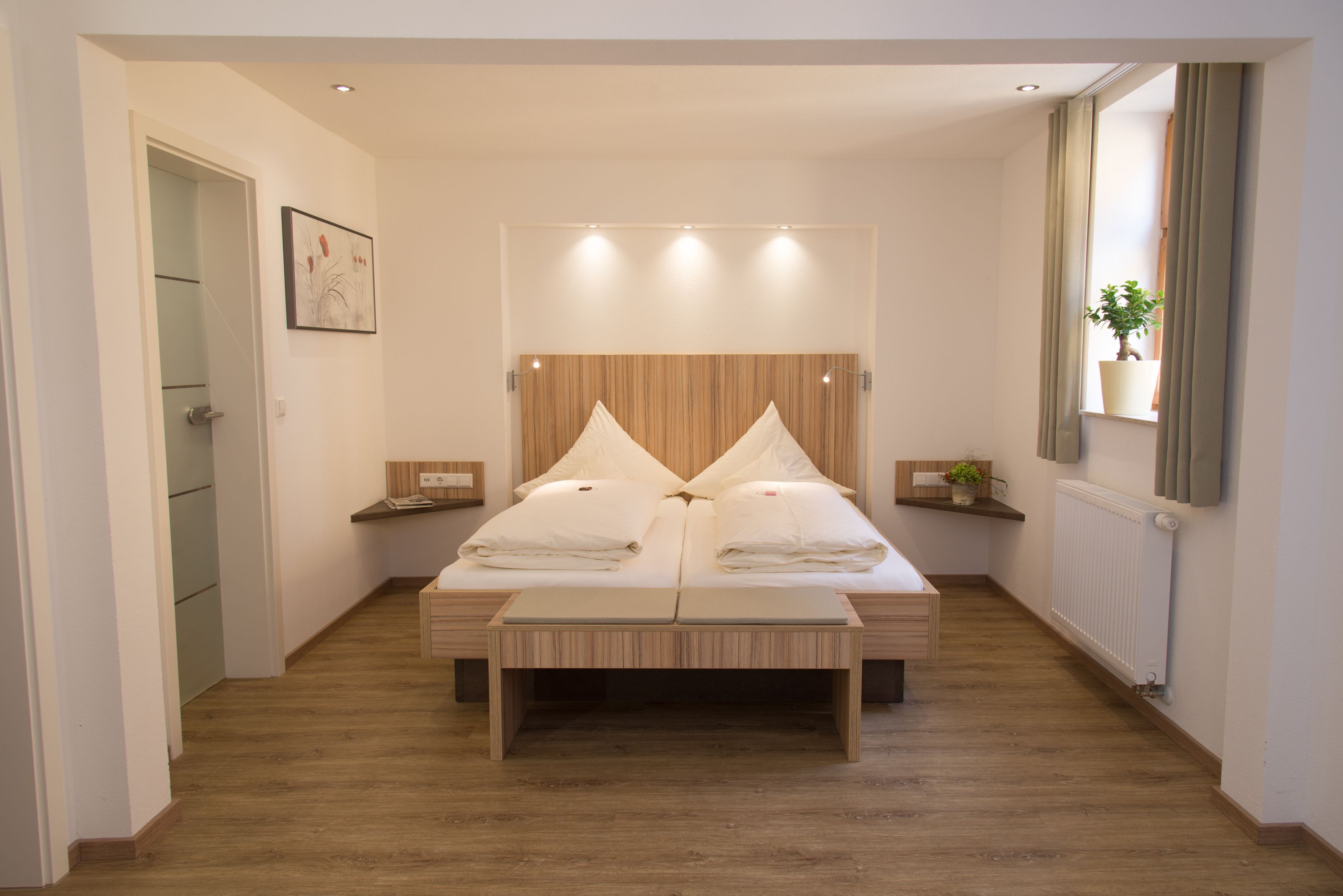Ein modernes Schlafzimmer mit einem Holz-Kopfteil, einem Doppelbett mit Kissen, einem Nachttisch mit Lampe und einem Blumentopf am Fenster.