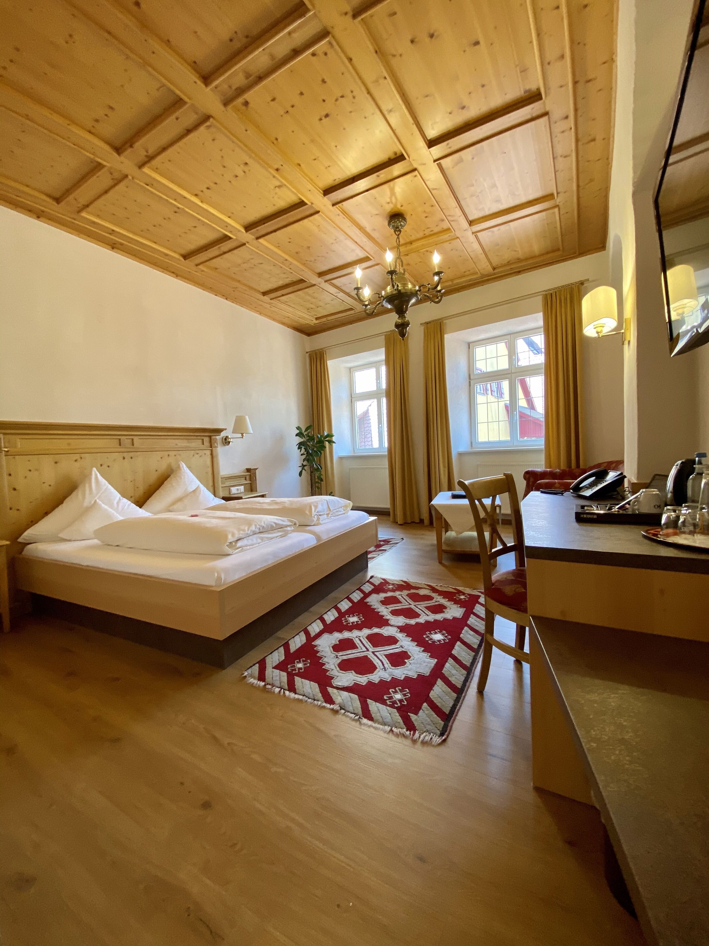 Ein Hotelzimmer mit Holzbalkendecke, einem Bett mit weißen Leinen, einem Schreibtisch mit Stuhl und einem roten Teppich mit Muster.
