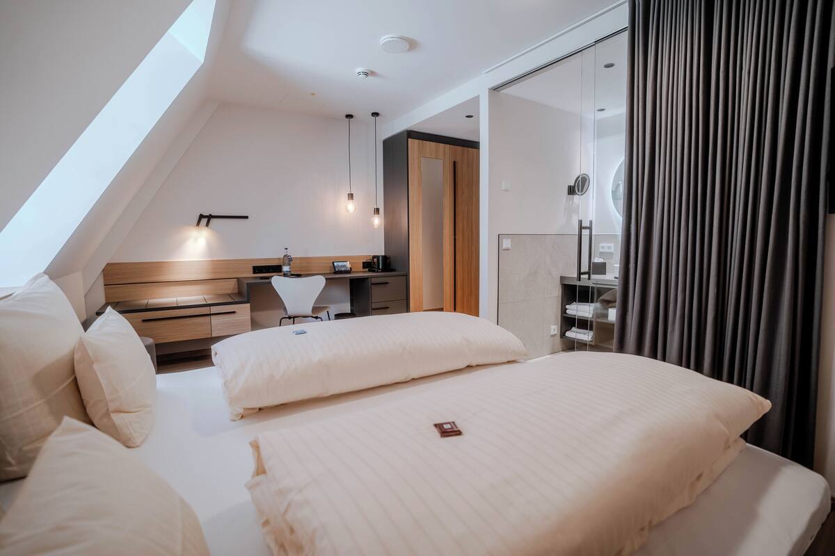A modern hotel room with a sloped ceiling, a desk with a chair, a bed with white linens, and a large mirror.