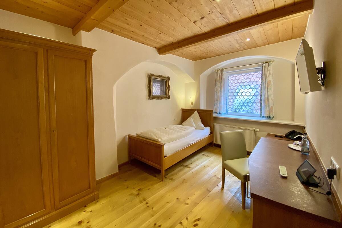 A bedroom with a wooden bed, a chair, a desk with a computer, a window with curtains, and a radiator.