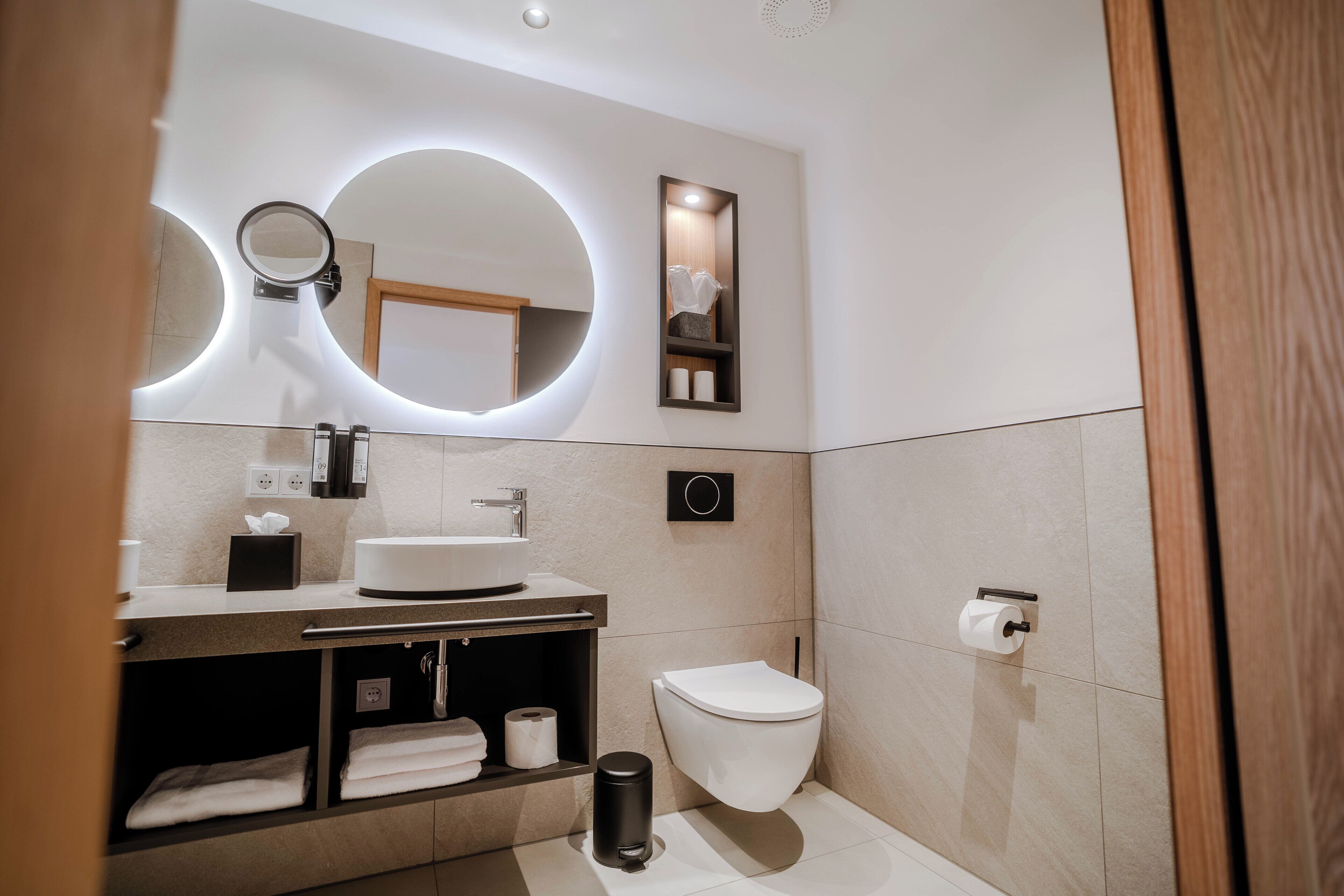 A modern bathroom with a round mirror, a sink, a toilet, and a trash bin.