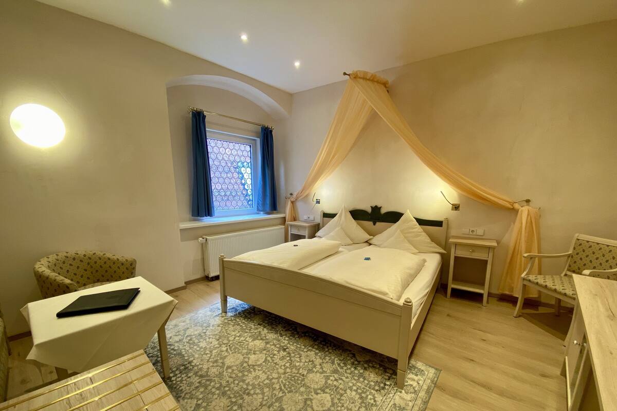 A bedroom with a bed, a chair, a small table, a window with curtains, a radiator, and a ceiling light.
