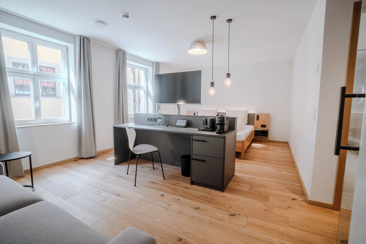 A modern, compact living space with a wooden floor, a grey sofa, a desk with a chair, a bed, and a TV mounted on the wall.