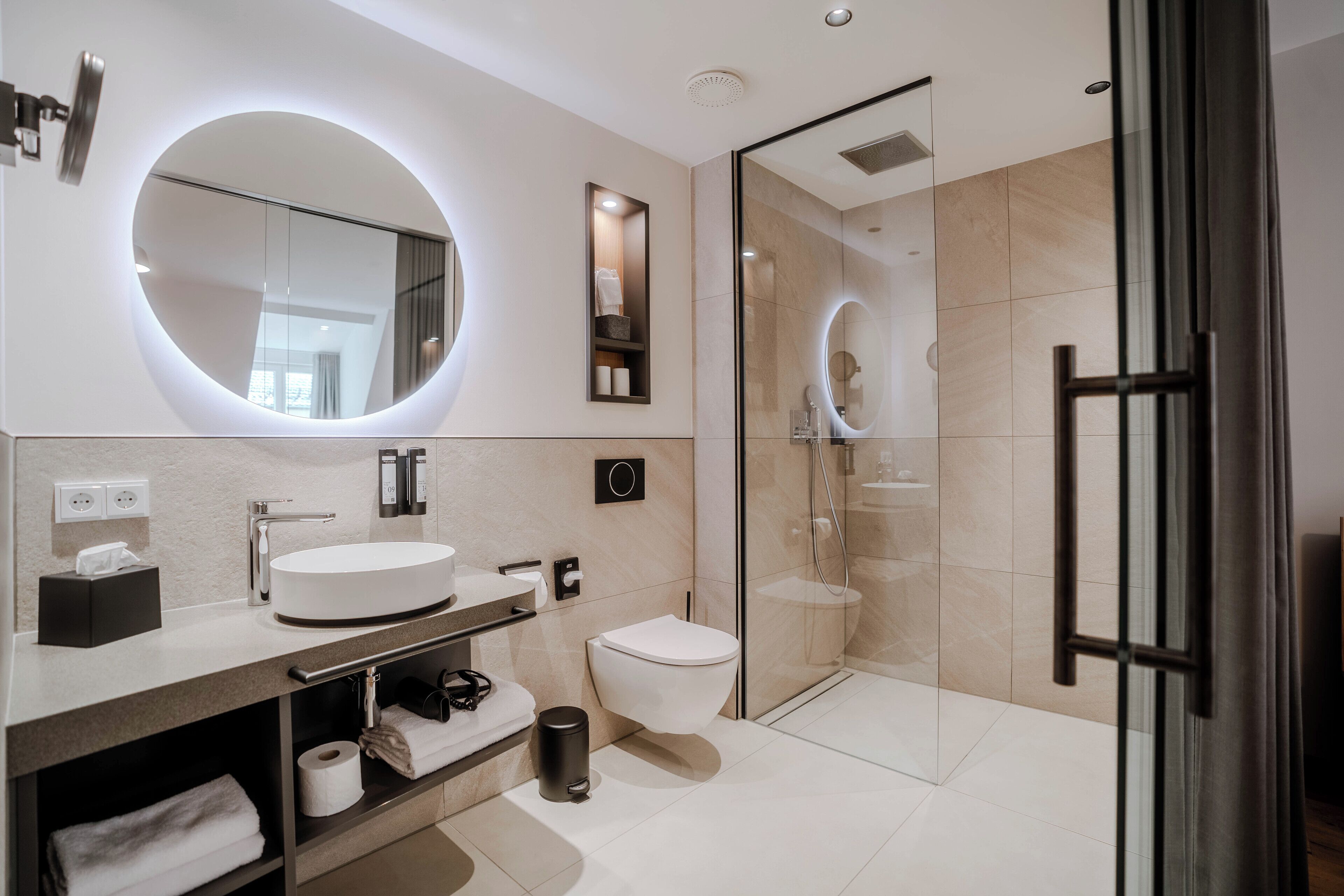 A modern bathroom with a round mirror, a white sink, a toilet, and a glass-enclosed shower.