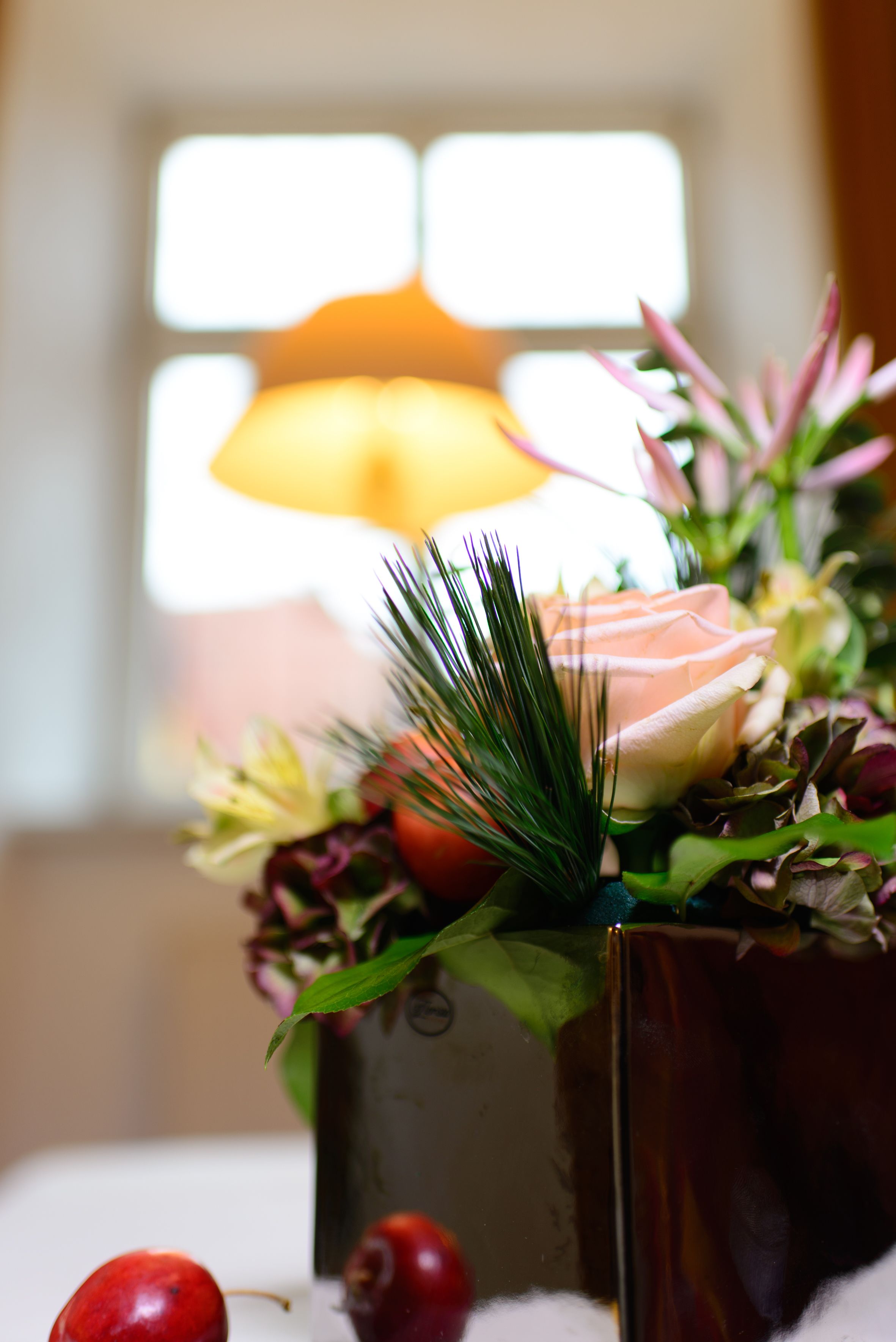 A close-up of a floral arrangement in a vase, with a blurred background featuring a window and a lamp.