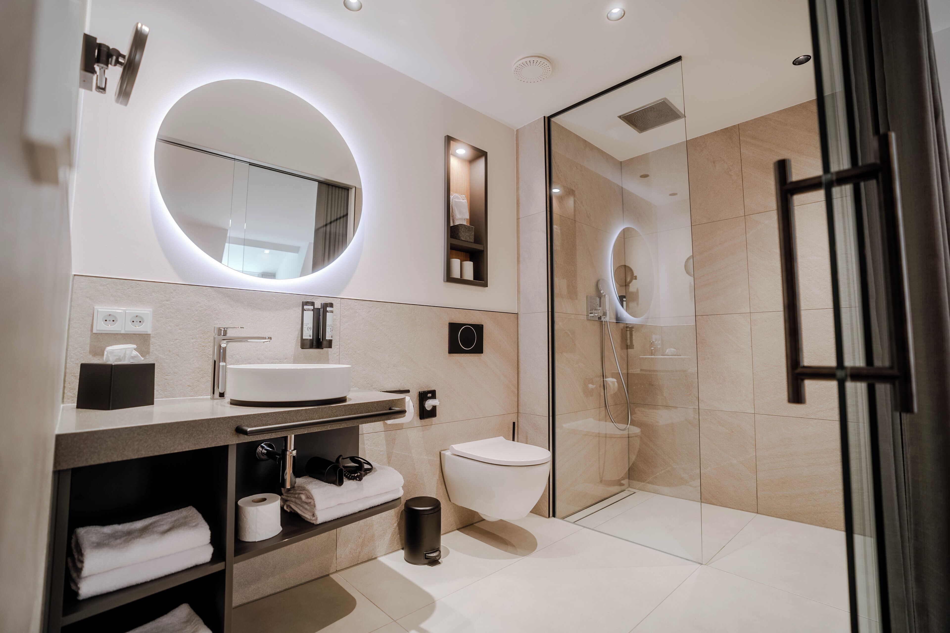A modern bathroom with a round mirror, a toilet, a glass-enclosed shower, and a vanity with a sink and towel storage.