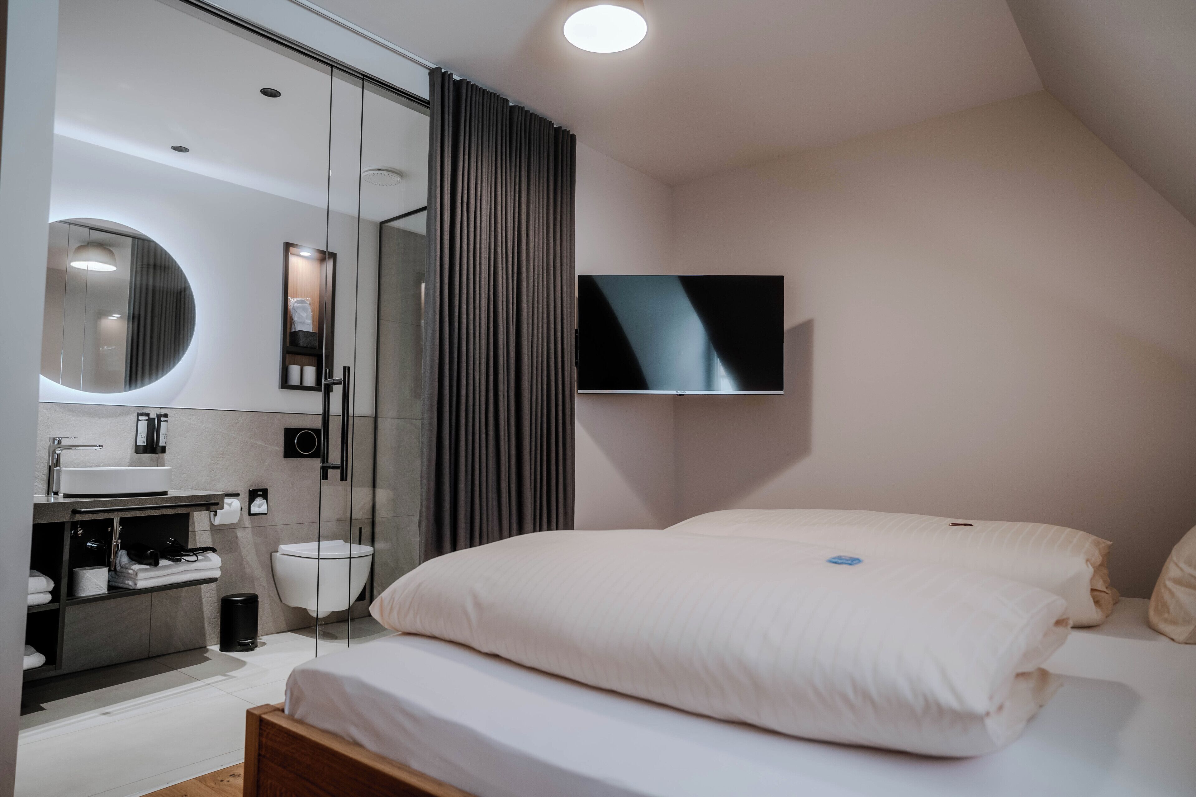 A modern hotel room with a large bed, a TV mounted on the wall, a bathroom with a mirror, and a shower.