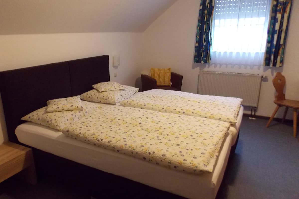 Ein Doppelzimmer mit zwei Betten, einem kleinen Holfnachtkästchen, einem Stuhl und einem Heizkörper.