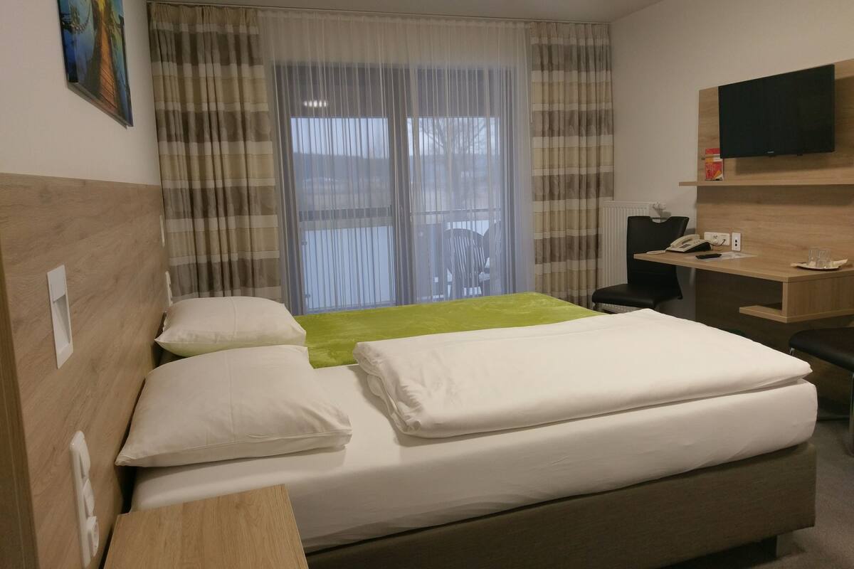 Ein Hotelzimmer mit Bett, Schreibtisch, Stuhl, Fernseher und einem Fenster mit Vorhängen.