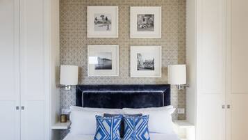 A neatly arranged bedroom with a patterned wallpaper, a dark headboard, and four framed photographs on the wall.