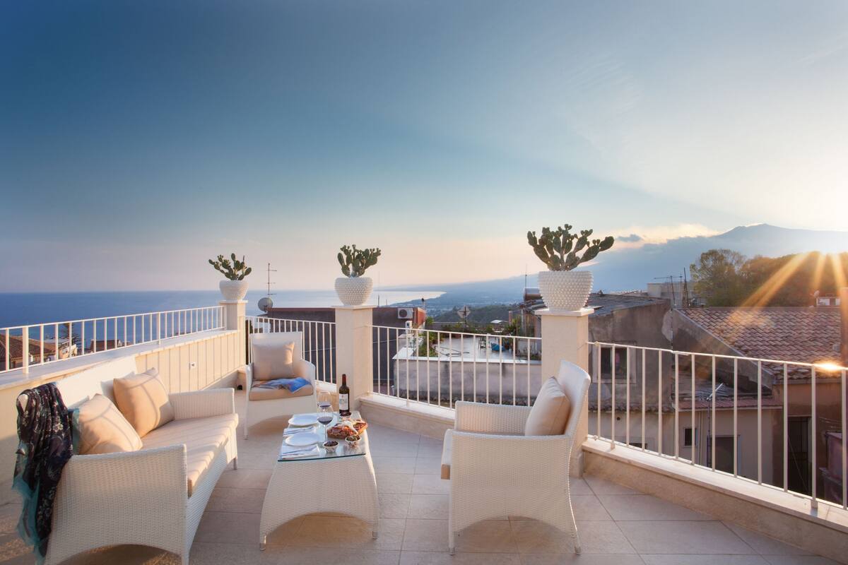 Une terrasse dotée de meubles en osier blanc, une table dressée avec des assiettes et des verres, et des jardinières contenant des cactus.