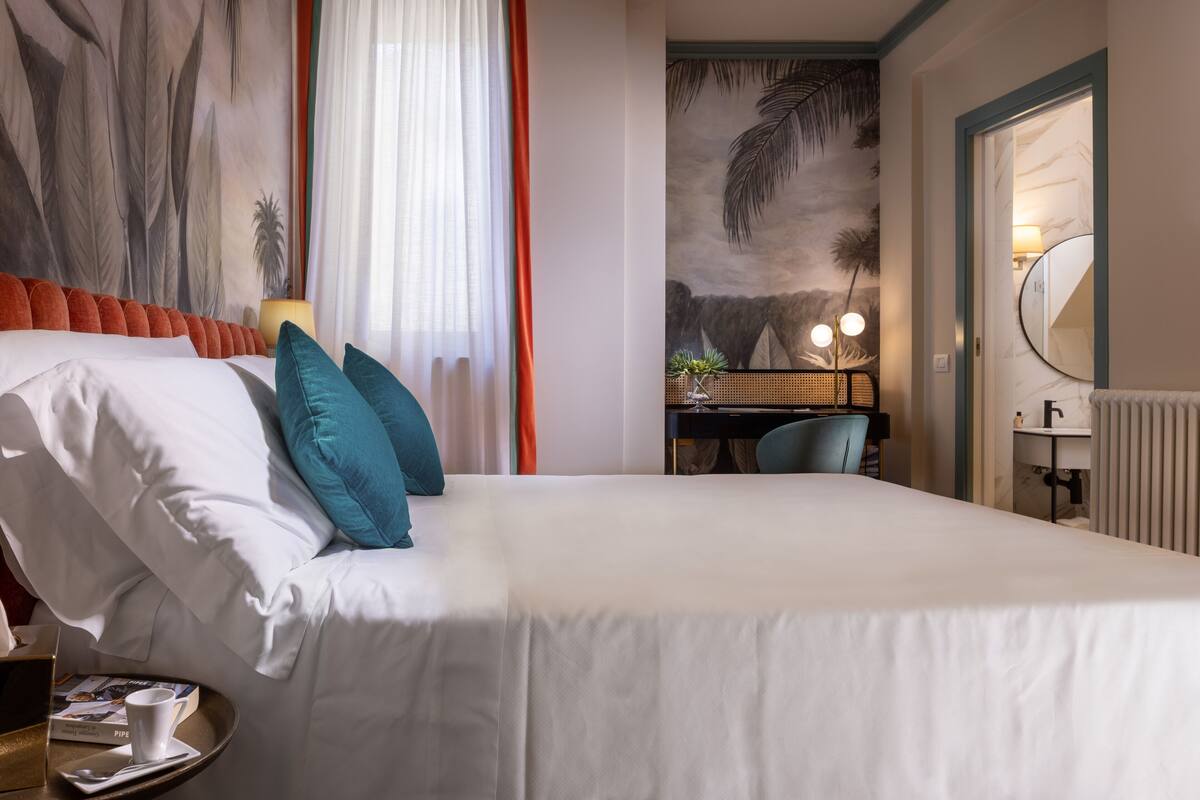 Une chambre d’hôtel avec un grand lit, un bureau avec une chaise, une petite table avec un plateau et une tasse, et une grande fresque murale.