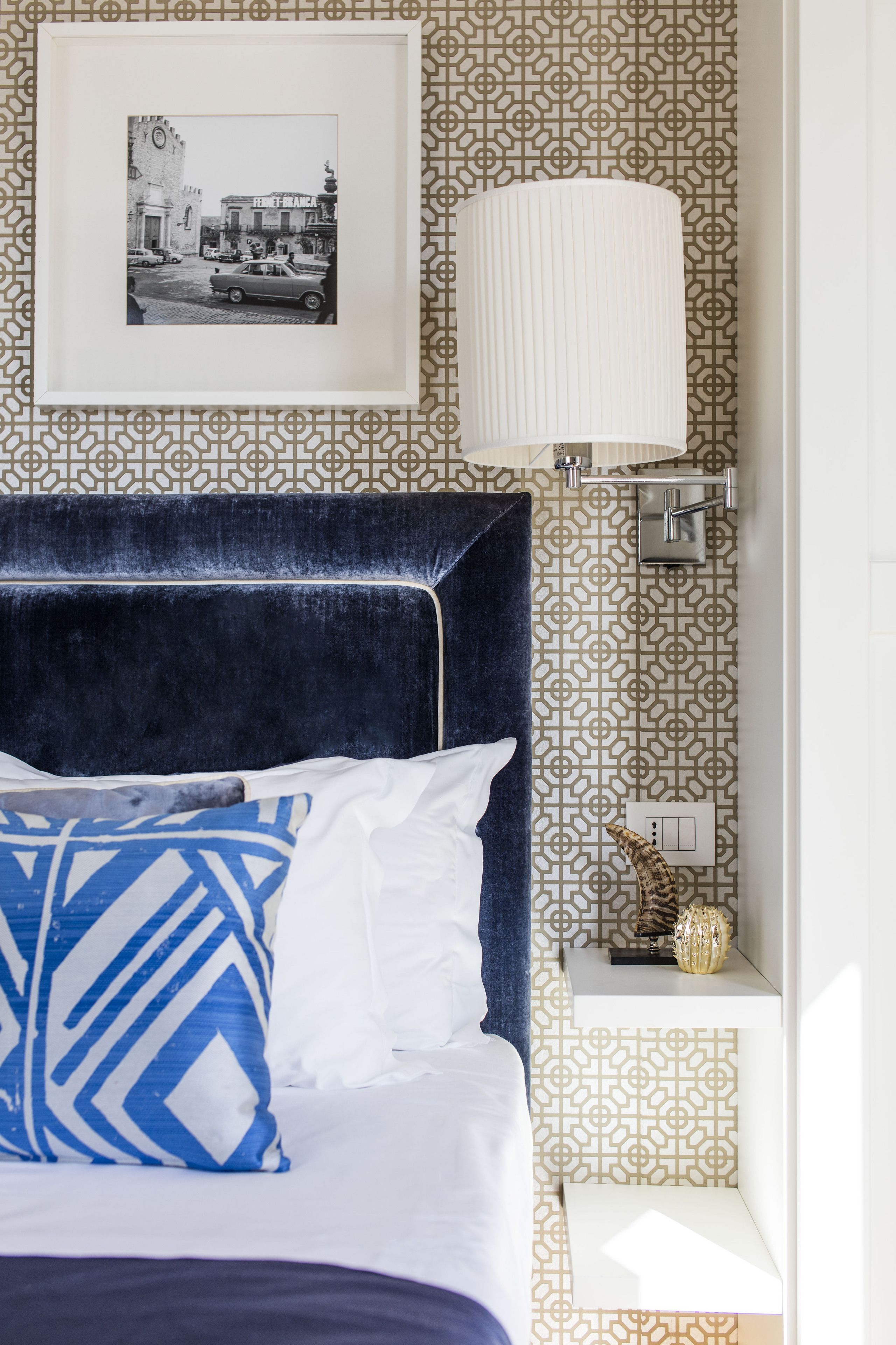 A neatly made bed with a blue and white pillow, a framed picture above it, and a patterned wall.