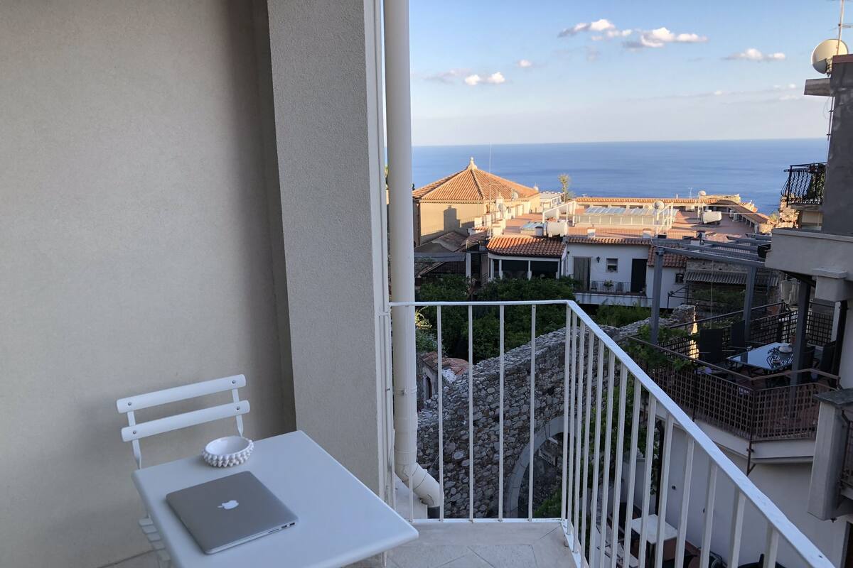 Un balcon avec une table et une chaise blanches, donnant sur une ville côtière avec ses bâtiments et la mer.