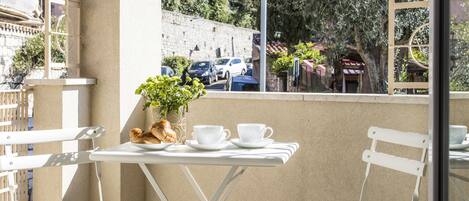 A balcony with a table and chairs, a potted plant, and a view of the street and trees.