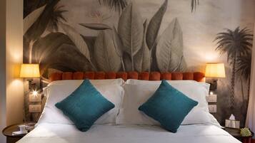 A hotel room with a bed, two teal pillows, a headboard with orange accents, and a mural of tropical leaves on the wall.